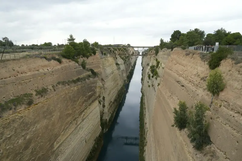 The Corinth Canal cut through pale rock, seen from the bridge above, with a railway bridge and a viewing platform further along the channel