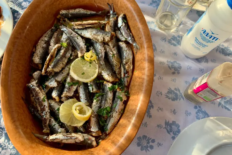 A bowl of fried small fish garnished with parsley and lemon slices on a taverna table