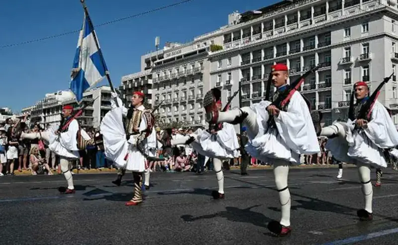 Evzones in ceremonial white uniform marching in a parade past a crowd on an Athens avenue