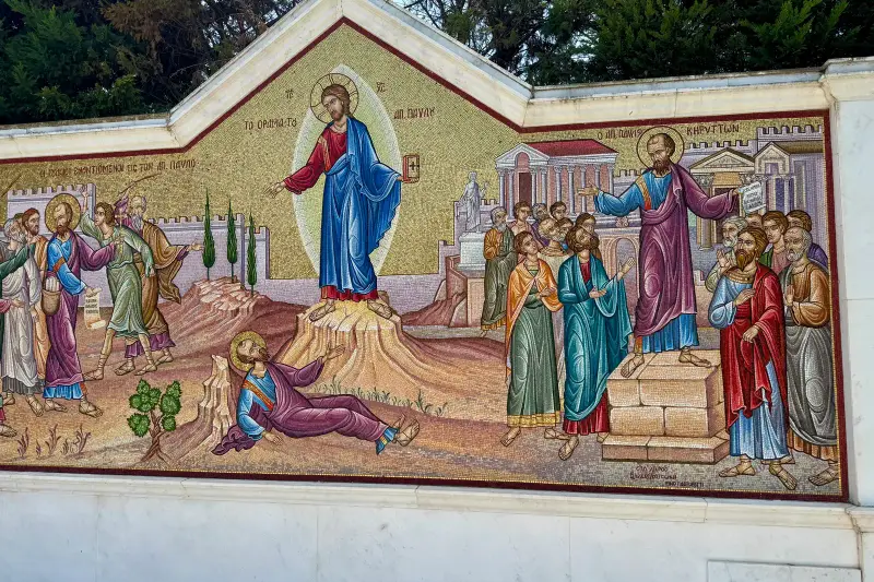 A large outdoor mosaic in two panels — on the left Christ appears in a halo of light to Saint Paul, who has fallen to the ground; on the right Saint Paul stands on a stone plinth preaching to a crowd